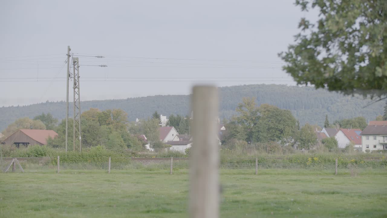 locomotora eléctrica moderna roja con rayas blancas, capturada en movimiento por una cámara de mano, moviéndose de izquierda a derecha a través de un paisaje verde y rural, bajo un telón de fondo de árboles y colinas distantes