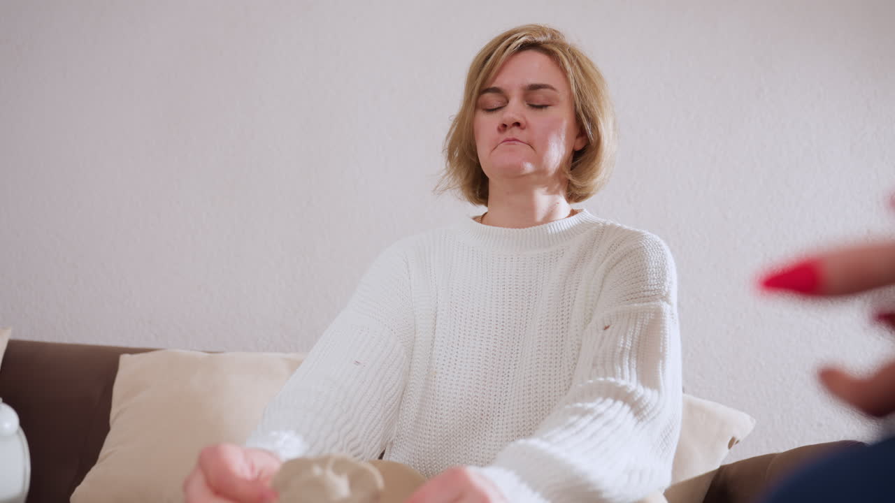 Healing seeker seated on sofa with eyes closed as she molds soft material during therapy session, engaging in deep emotional reflection while attentively listening to therapist in indoor environment