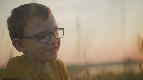 a young boy wearing glasses and a yellow shirt is sitting in a grassy field, smiling warmly. the scene captures a peaceful moment as someone in the background seems to engage with him