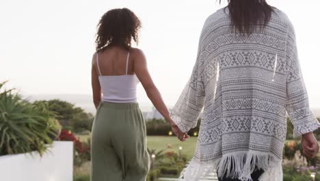 Rear-view-of-happy-biracial-lesbian-couple-holding-hands-and-walking-in-garden,-slow-motion