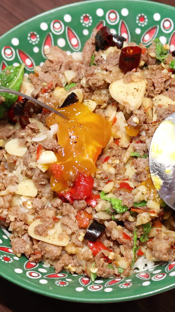 Una forchetta e un cucchiaio rompono e mescolano un tuorlo d'uovo liquido in un colorato piatto di riso tailandese con carne macinata, sotto un'illuminazione brillante e uniforme, in primo piano.