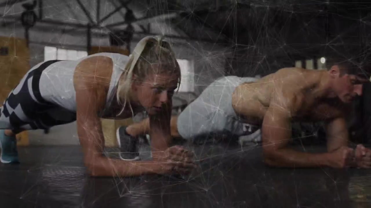 Atleta donna in plank mentre il partner uomo si abbassa, entrambi che rafforzano il core sul pavimento della palestra.