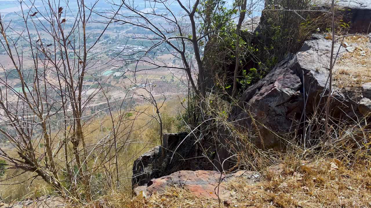 Person records vast countryside valley from high rocky cliff viewpoint under bright midday sky