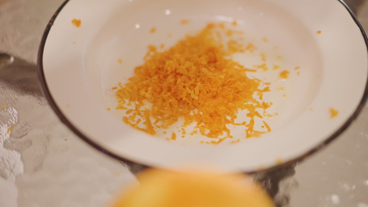 Grated orange zest piled in bowl, enamel plate on glass table with soft warm lighting, fine citrus shavings gathered into a small heap, detailed texture of peel, careful pastry prep for dessert
