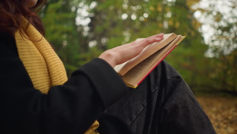 close-up of woman with red wavy hair wearing yellow beret and sunglasses flipping through book in her hand, face slightly blurred in background, autumn leaves scattered around