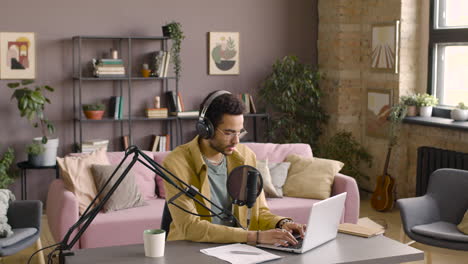 man wearing headphones while he is talking into a microphone, typewriting on laptop and recording a podcast