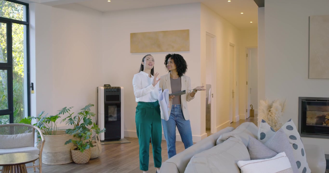 Mulheres diversas entrando na sala de estar de blazers, comparando planos com o espaço usando um tablet, avaliando.