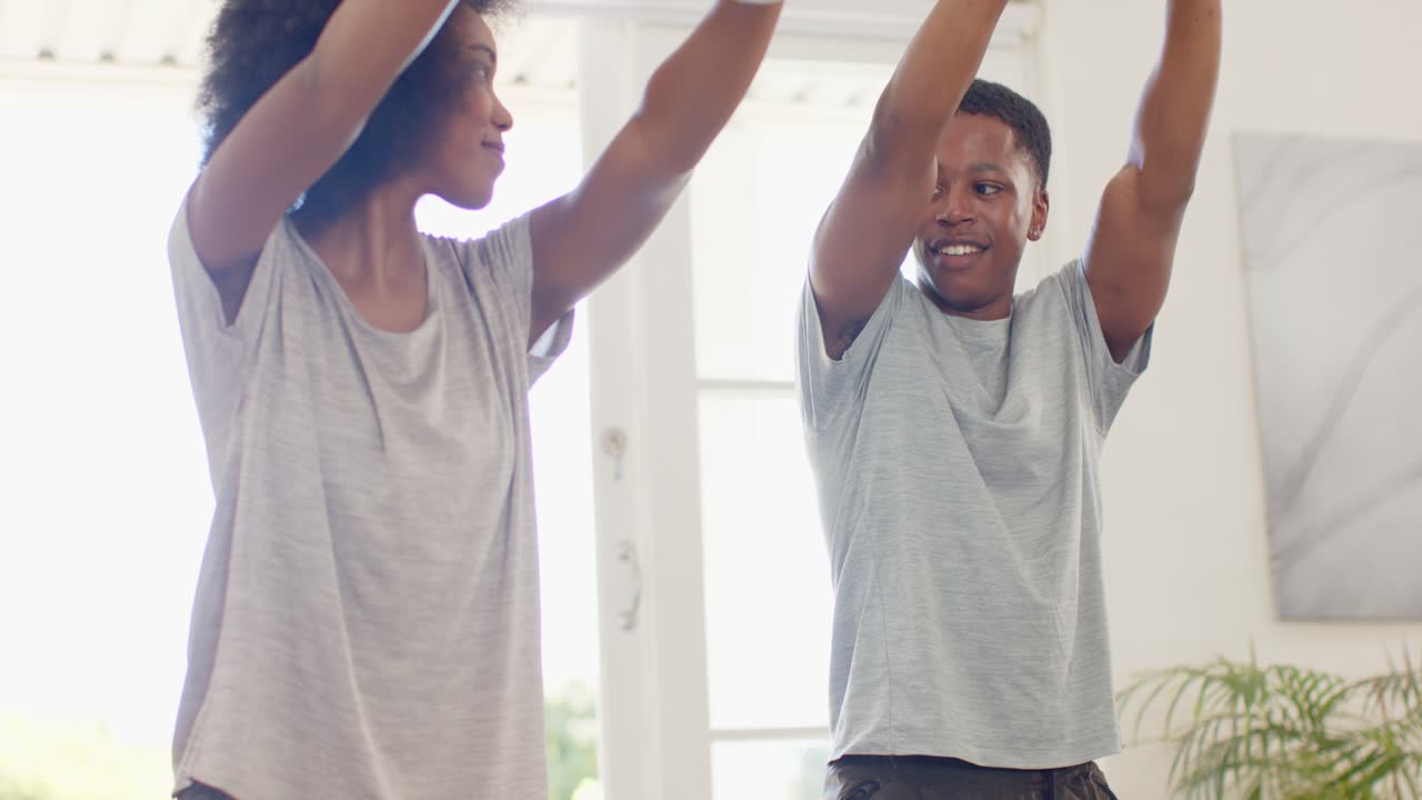 African American couple kneeling on mats at home, warming up, stretching overhead with water bottle