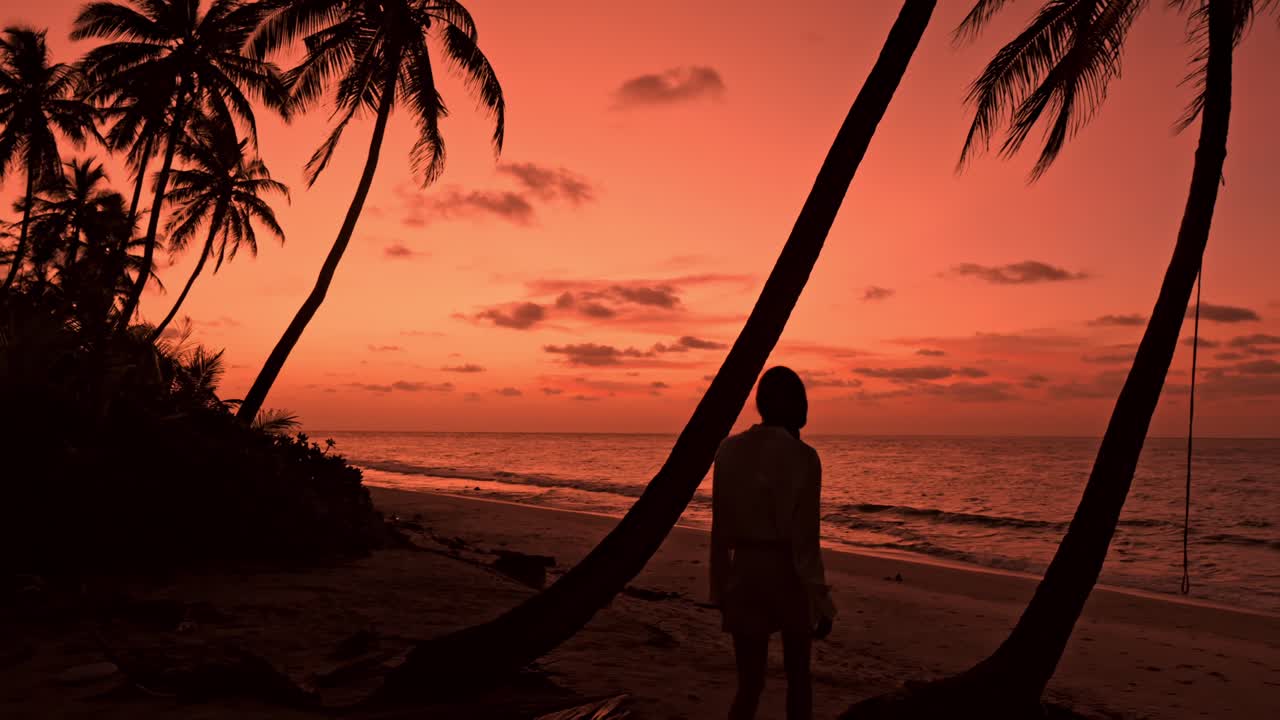 A breathtaking view captures a lone woman strolling along a pristine, palm-lined beach in Fuvahmulah, Maldives at sunset.