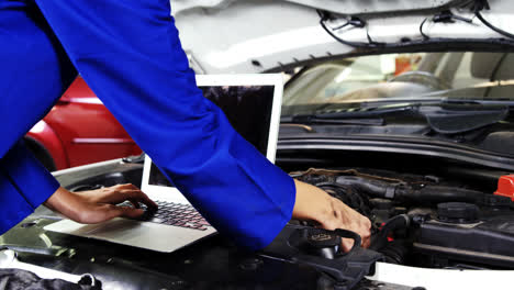 mecánica femenina usando una computadora portátil mientras hace servicio al motor de un automóvil