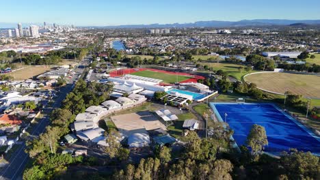 drone footage captures a vibrant sports park with fields, pools, and surrounding cityscape under clear skies