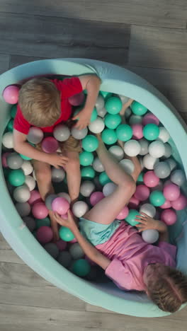 cheerful male toddler and female preschooler play together lying in dry pool. smiling girl throws plastic balls to brother and toys roll on floor