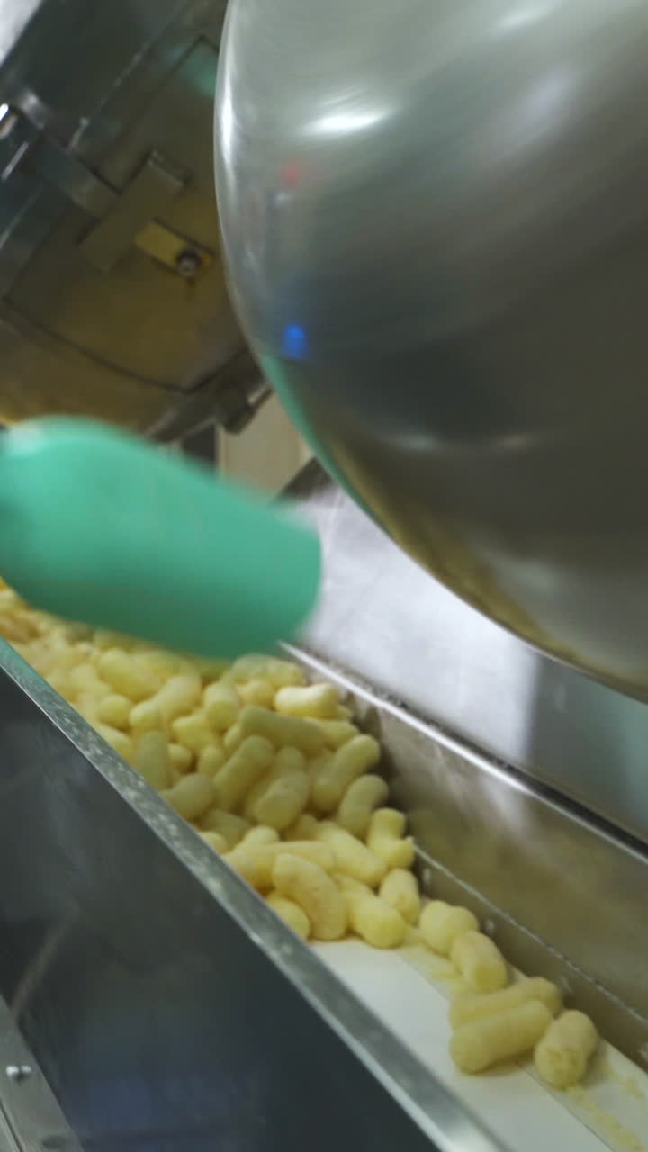 Pouring corn sticks of snacks from a metal tank onto an automatic conveyor belt. Hand of a worker with a shovel. Food factory. Vertical video