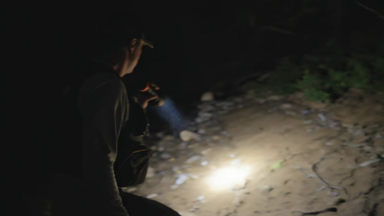 Lifting flashlight man in pocketed vest and cap scanning ground, walking uneven bank, copy space