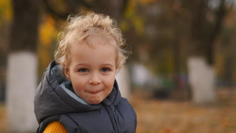 closeup portrait of little child with blonde curly hair in autumn park in city funny face and grimacing of baby boy walking children