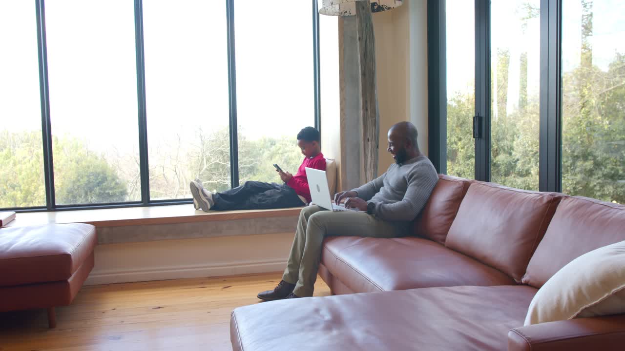 African American father and son at home, father starting work on laptop while son scrolling phone