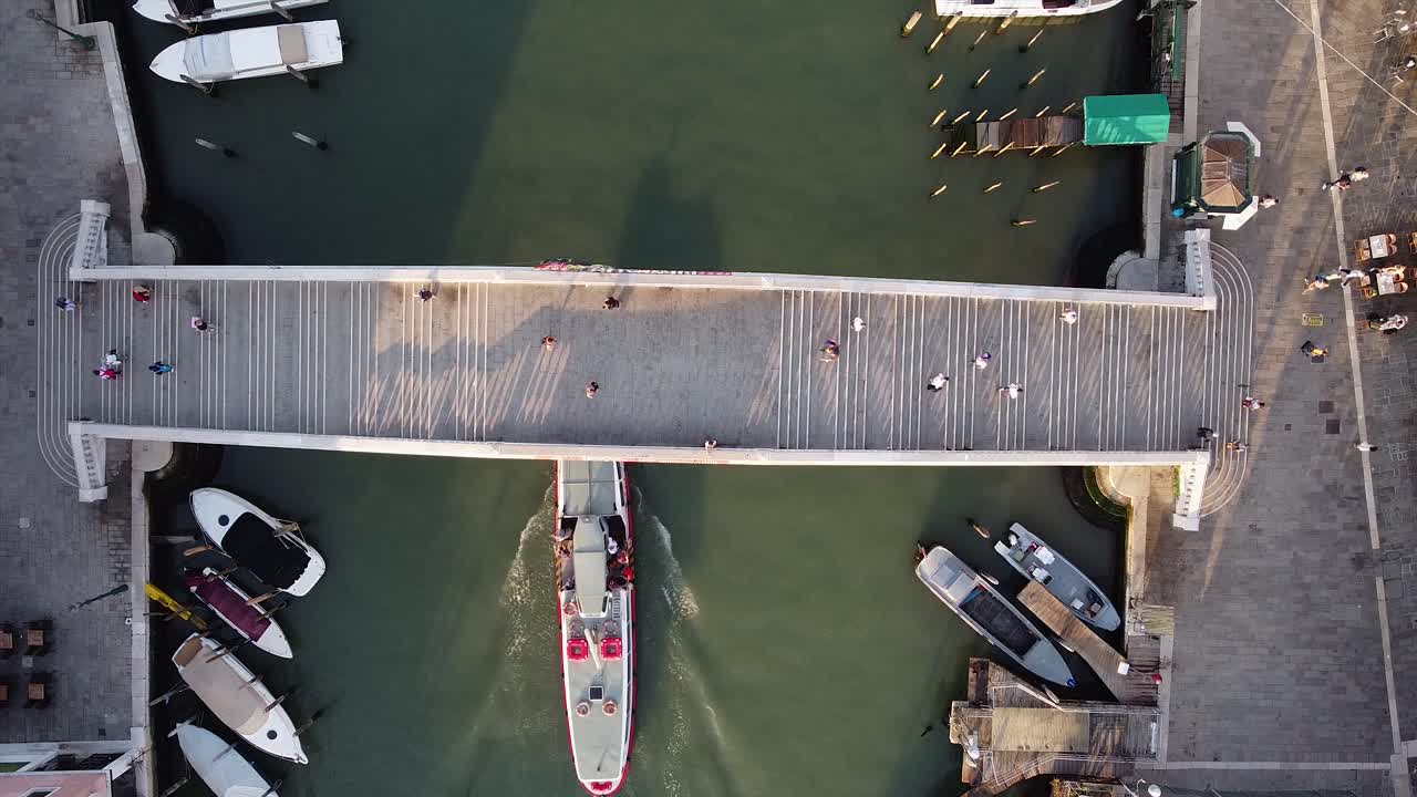 Top-down aerial view of tourists walking over a footbridge in Venice, Italy, as boats pass in the canal underneath