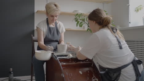 A female potter&#039;s wheel sculpting teacher explains how to work and teaches an elderly woman to work with clay and make mugs and jugs. Master class for pensioners. Pottery courses
