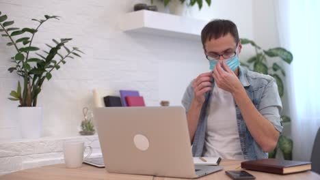 young caucasian man stay home working from home wearing protective mask using laptop and internet. cozy home office, workplace coronavirus pandemic, covid 19 quarantine. remote work,freelancer