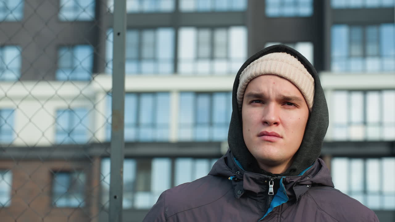 молодой взрослый в капюшоне и шапочке с мячом в правой руке проходит через барный забор с серьезным выражением лица, высокое офисное здание на заднем плане, и солнечный свет отражается на нем