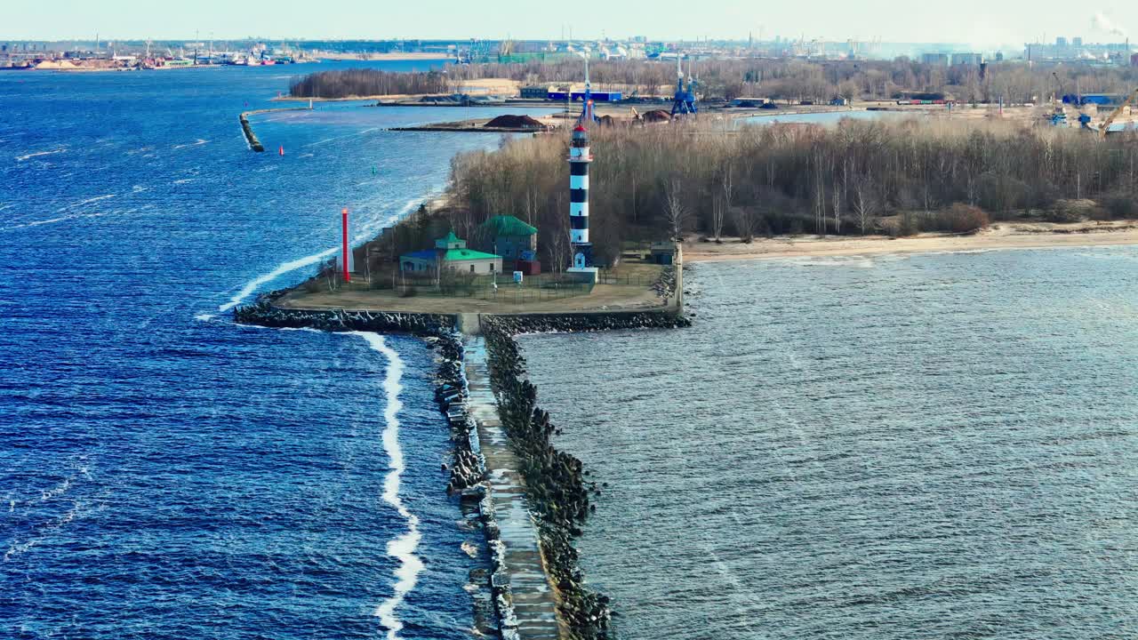 A black and white striped lighthouse stands at the end of a long jetty splitting bright blue Baltic Sea from calm estuary waters, bordered by forest and industrial skyline.