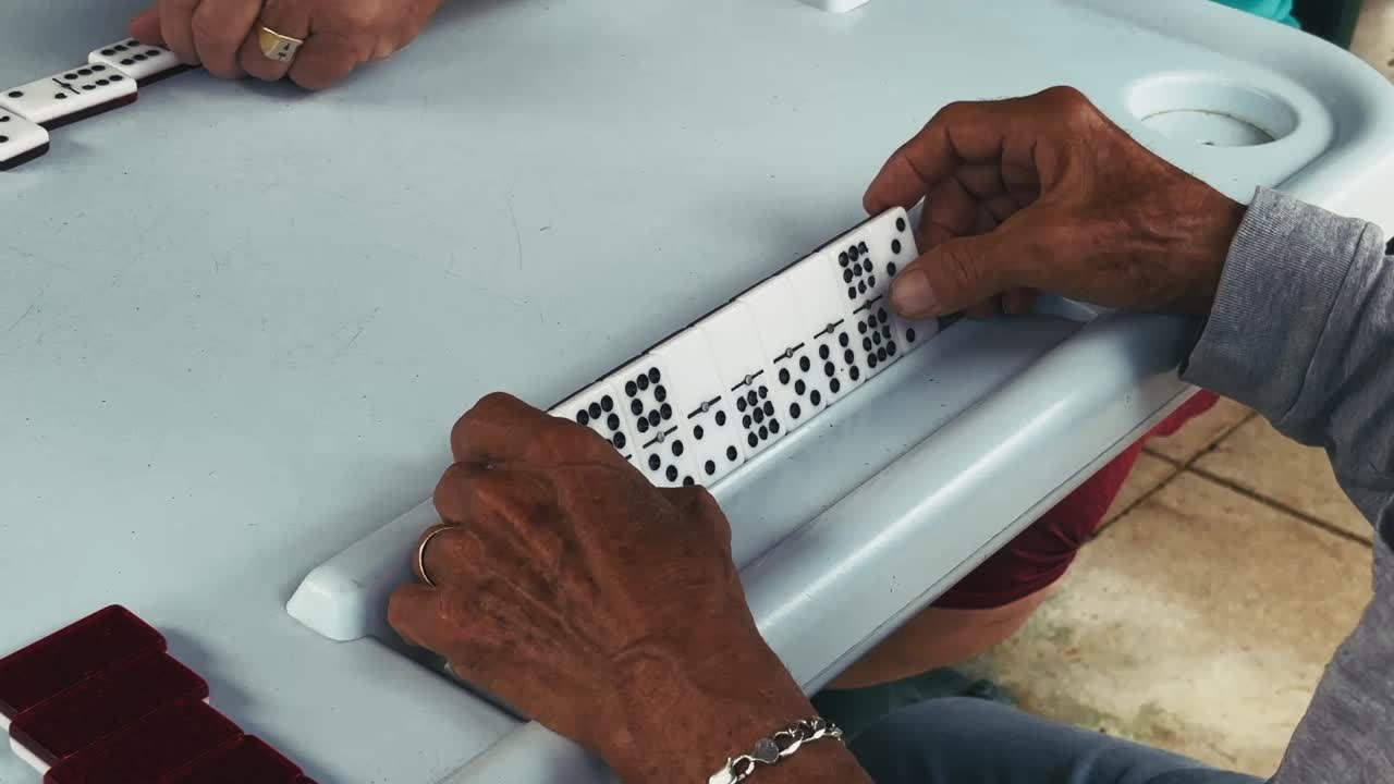 Hands grip a row of domino tiles on a plastic rack while playing a tabletop game at a light-colored table in Miami