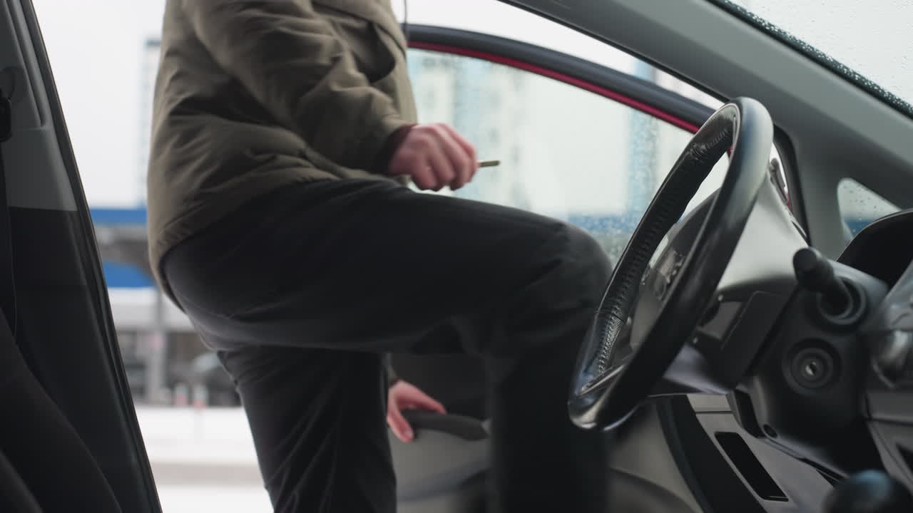 Close up interior view of car with steering wheel and door visible, water droplets on window, blurred urban background and man in winter jacket entering vehicle on cold snowy day