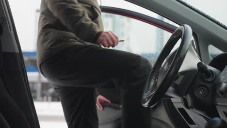 close up interior view of car with steering wheel and door visible, water droplets on window, blurred urban background and man in winter jacket entering vehicle on cold snowy day