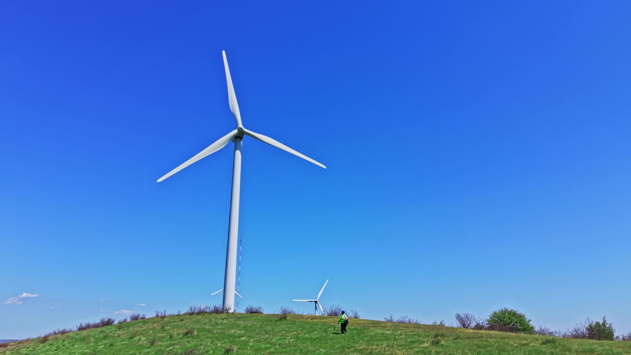 Engineer checks operation of windmills on a sunny day in the countryside while standing on a green hill under a clear blue sky with large turbines in view