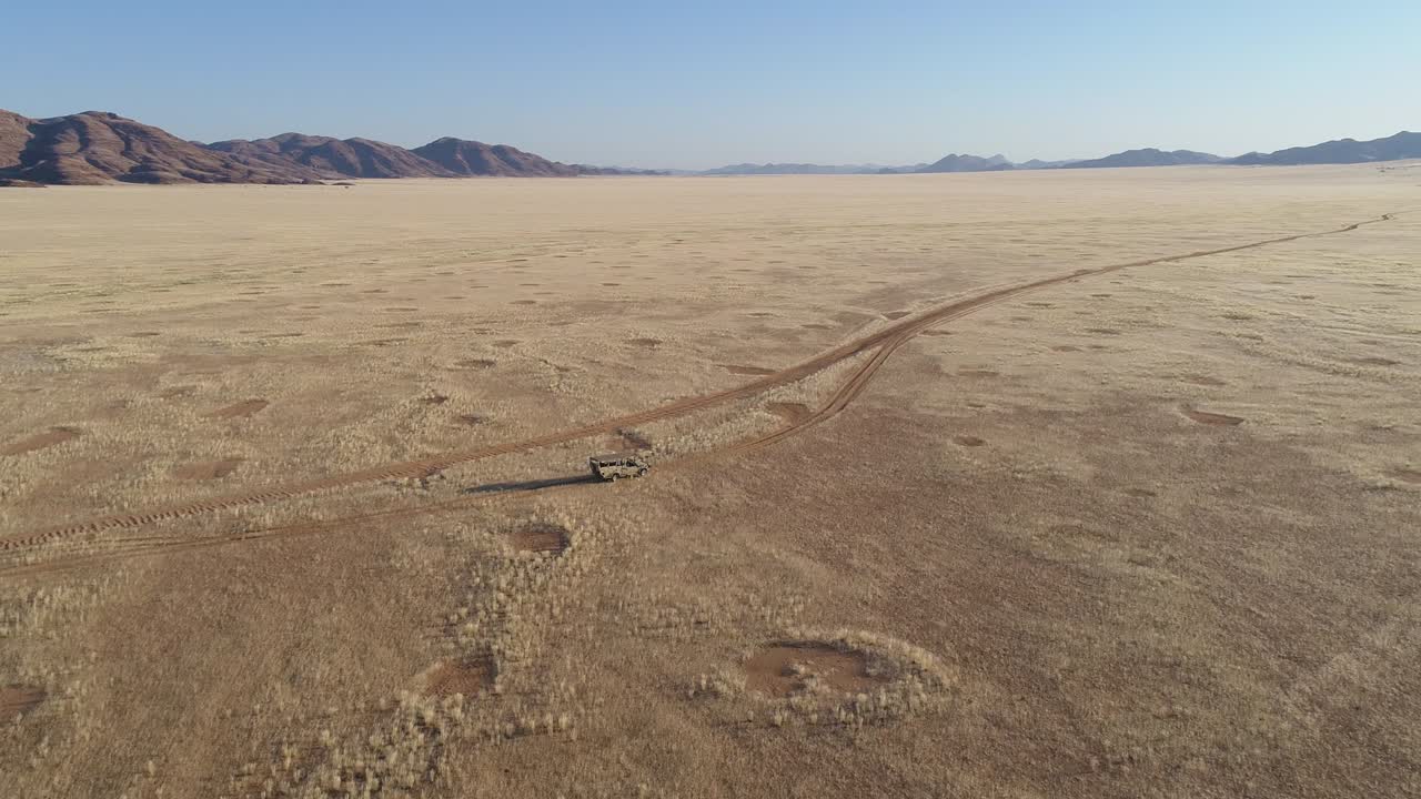 4K high tilt up aerial view of a 4x4 safari vehicle driving on a sand track through the African savannah grasslands with the famous fairy circles, Namibia