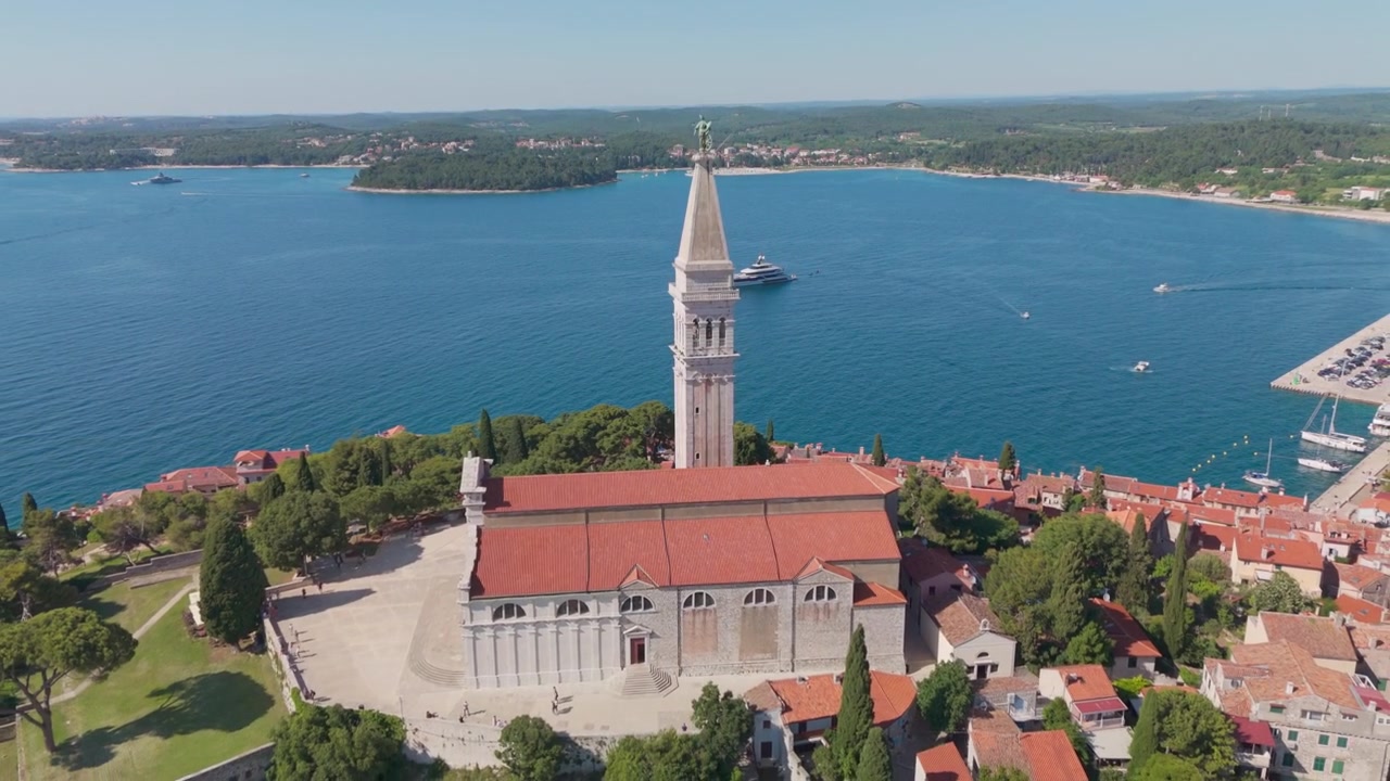 Drone ravvicinato sopra i tetti di Rovigno vicino a Sant'Eufemia, che rivela il campanile della chiesa in stile veneziano e vola all'indietro in un'ampia vista della penisola del centro storico nella luminosa luce adriatica.