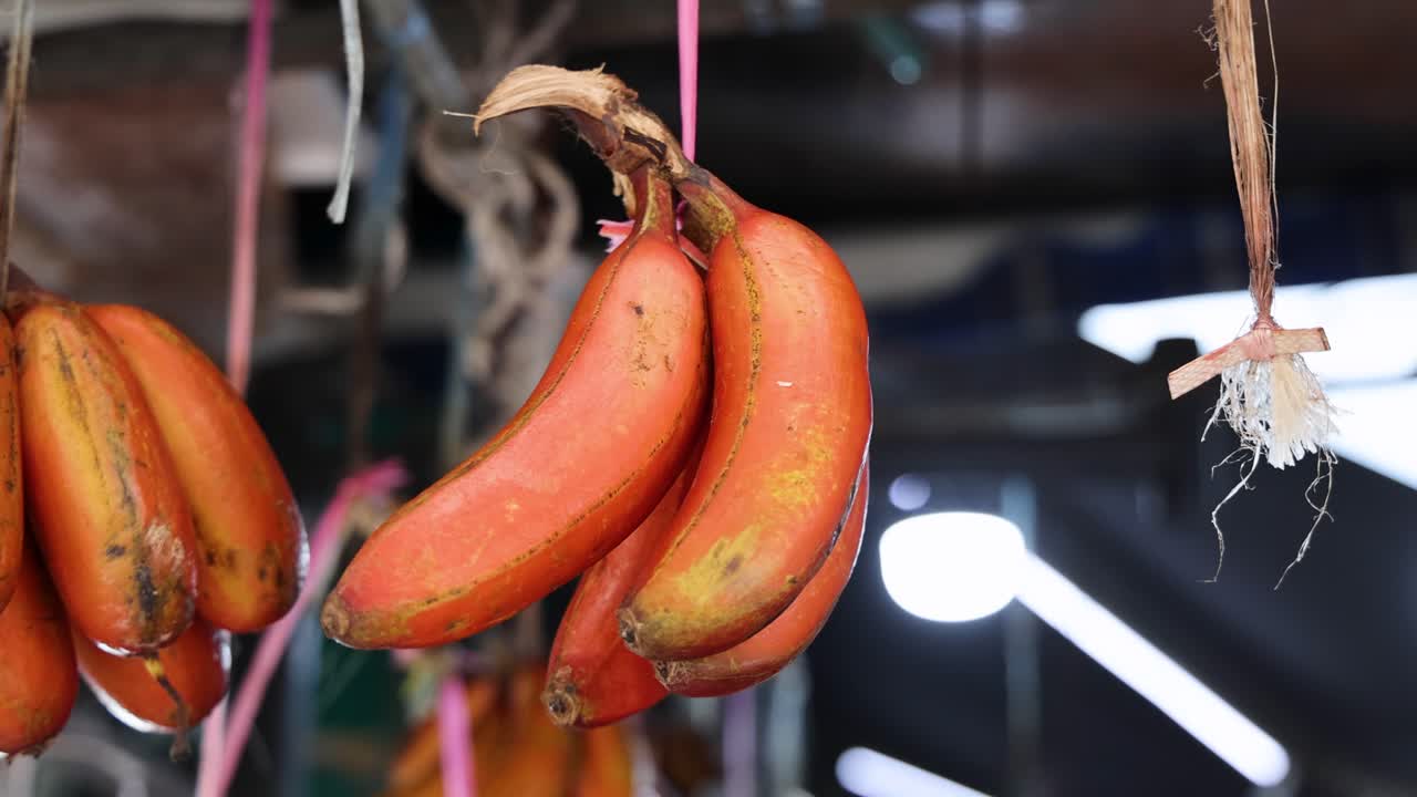 A bunch of ripe red bananas hangs from twine in an open-air market, with natural daylight and slight camera movement revealing a bustling Singapore setting