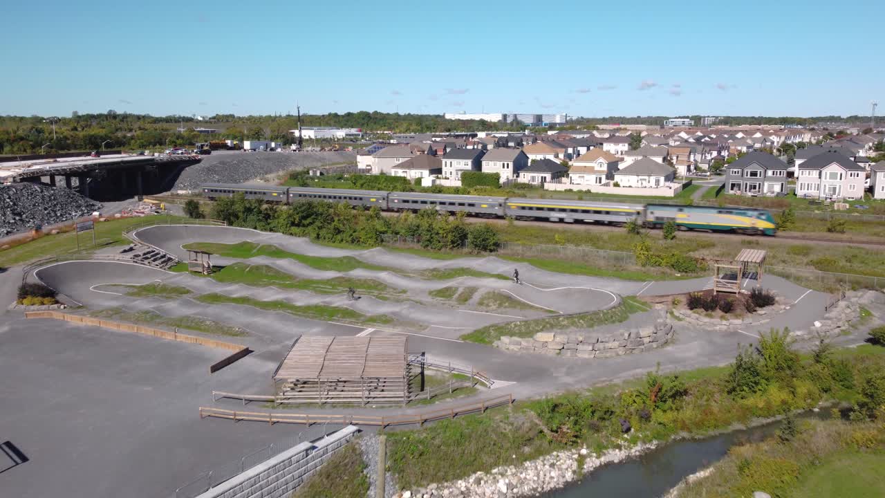 Luftfoto af en BMX-cykelpark på en solrig dag i forstæderne, mens to teenagere cykler, og et tog kører forbi.