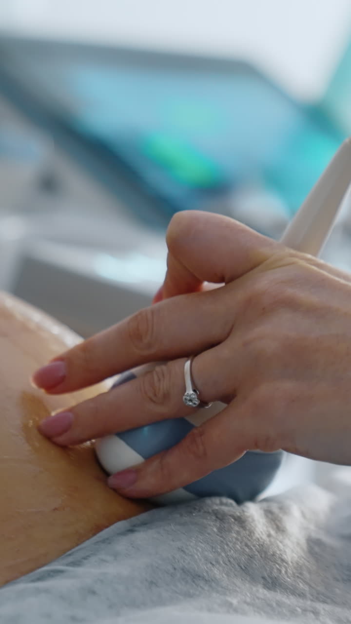 Female doctor's hand moving the device of ultrasound machine by the big pregnant belly covered with gel. Close up. Vertical video