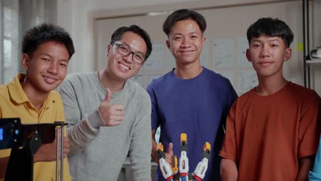 hombre asiático y los niños con una computadora portátil e impresión 3d sonriendo y mostrando los pulgares hacia arriba gesto a la cámara mientras discuten sobre una mano cyborg en la escuela