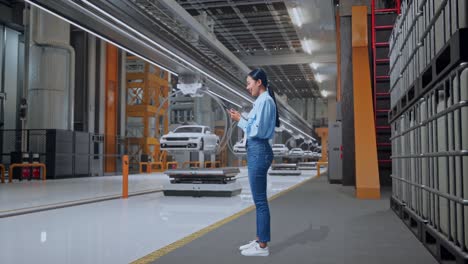 vista lateral completa del cuerpo de una trabajadora profesional asiática de pie con su teléfono en una fábrica de automóviles ev, instalación industrial de alta tecnología, trabajando continuamente con su teléfono