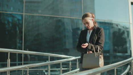 white lady preparing for lecture, standing near modern glass building, checking handbag to retrieve car keys, sunlight casting warm glow, building reflects her image and urban surroundings