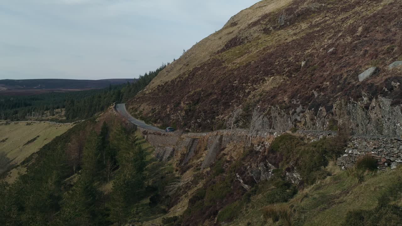 A car drives along a winding mountain road near Lough Tay, Wicklow, Ireland. The scenic route passes through rugged hills and forests, showcasing the beauty of Irish countryside travel.