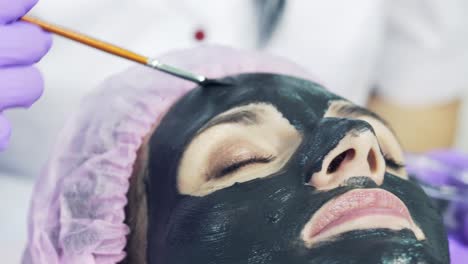 woman with closed eyes is getting facial cleansing mask by beautician at spa salon. apply black face mask, spa beauty treatment and skincare. closeup