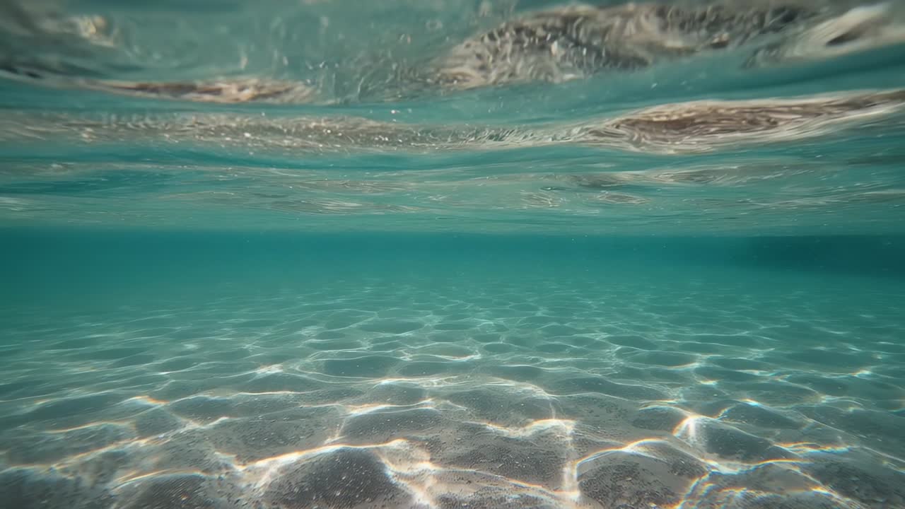 Telecamera che riprende un fondale sabbioso, il sole penetra nell'acqua creando bande di luce in movimento, catturando il movimento