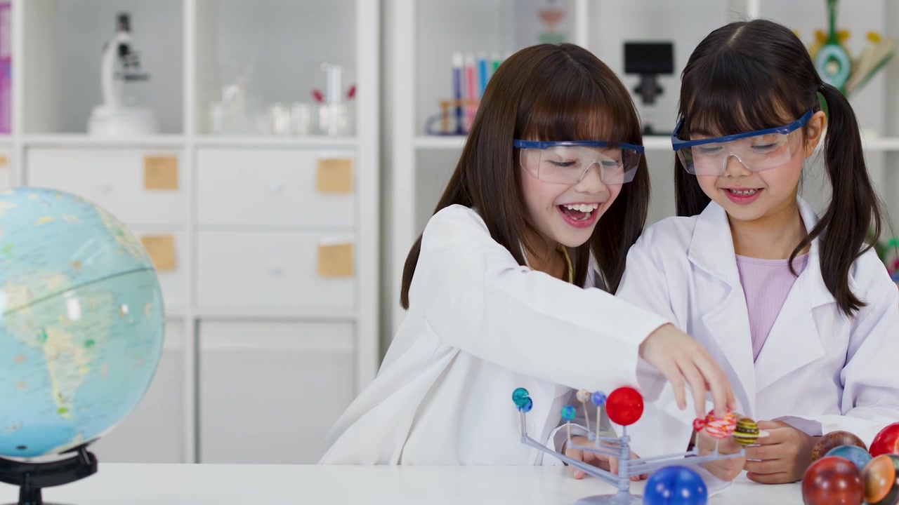 Two young students wearing safety goggles examine a planetary model in a bright classroom laboratory.