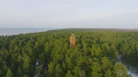 aerial view of bernati lighthouse surrounded by lush green pine tree forest with light snow, nordic woodland, baltic sea coast, sunny winter day, latvia, distant wide orbiting drone shot