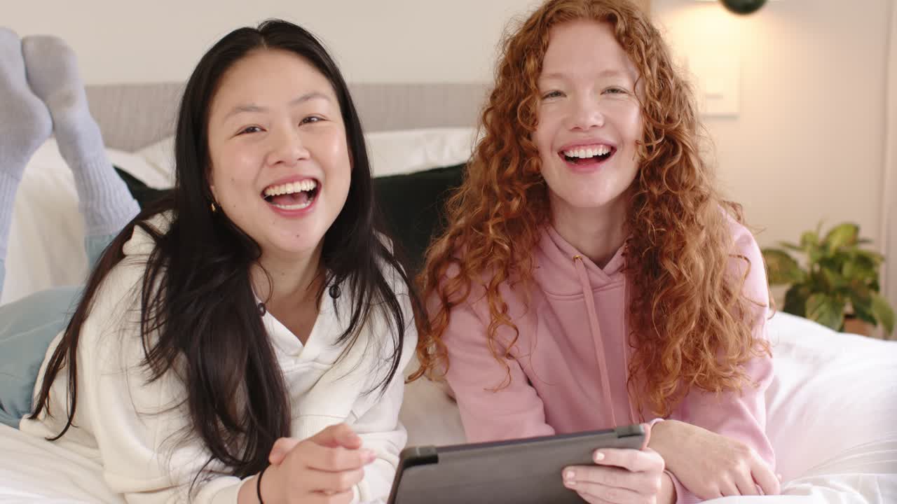 Diverse amiche distese sul letto che guardano un clip divertente su un tablet e sorridono alla fotocamera