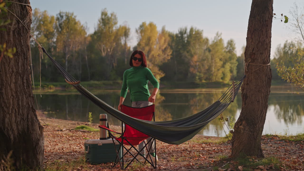 Donna bianca seduta in amaca in riva al lago che sistema frigo portatile e sedia, poi si alza per piegare l'amaca lasciando l'amaca vuota accanto al lago calmo con alberi autunnali che si riflettono nell'acqua, campeggiatrice solitaria che si gode la pace.