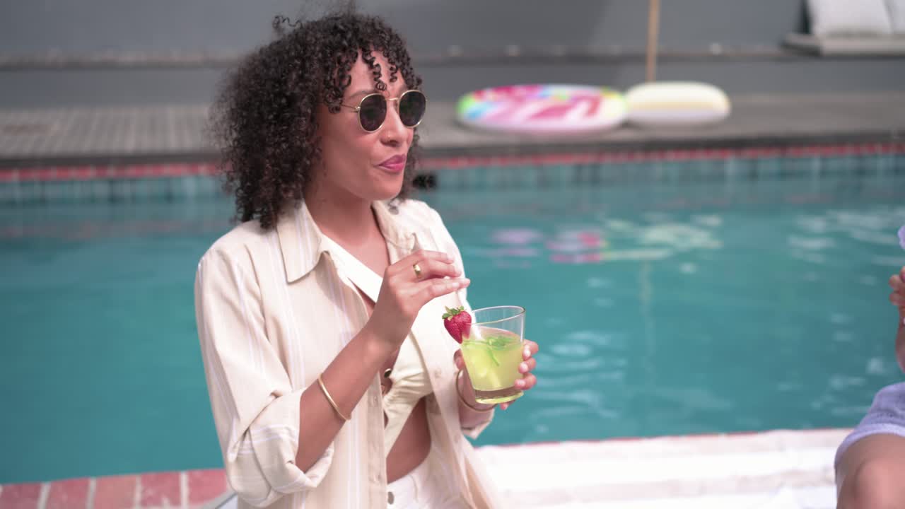 Verschiedene Freundinnen sitzen auf einem gestreiften Handtuch am Pool, die rechte Frau hebt einen Cocktail zum Anstoßen.