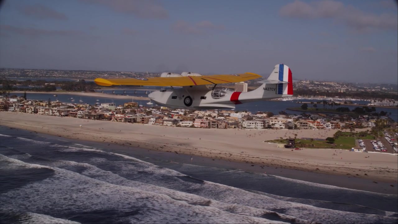Spektakulär drönarbild från luften av en historisk PBY Catalina flygbåt från andra världskriget som flyger över San Diegos kustlinje, med det ikoniska Hotel del Coronado och stadens silhuett synliga i bakgrunden.