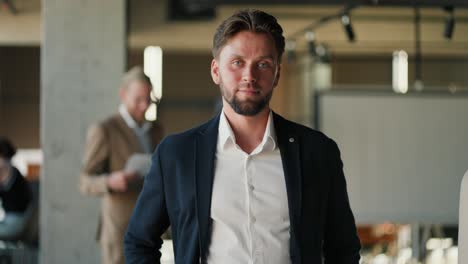 a young brunette office worker guy with stubble in a blue jacket and white shirt poses in a modern office against the background of his colleagues