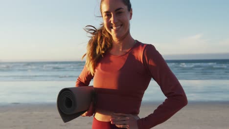 Porträt-Einer-Kaukasischen-Frau,-Die-Lächelnd-Eine-Yogamatte-Am-Strand-Hält