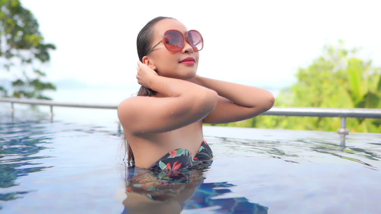 joven morena asiática de pie dentro de la piscina con un bikini florido, tocándose el pelo mojado levantando la cara con deleite y abriendo los ojos en bali, a cámara lenta