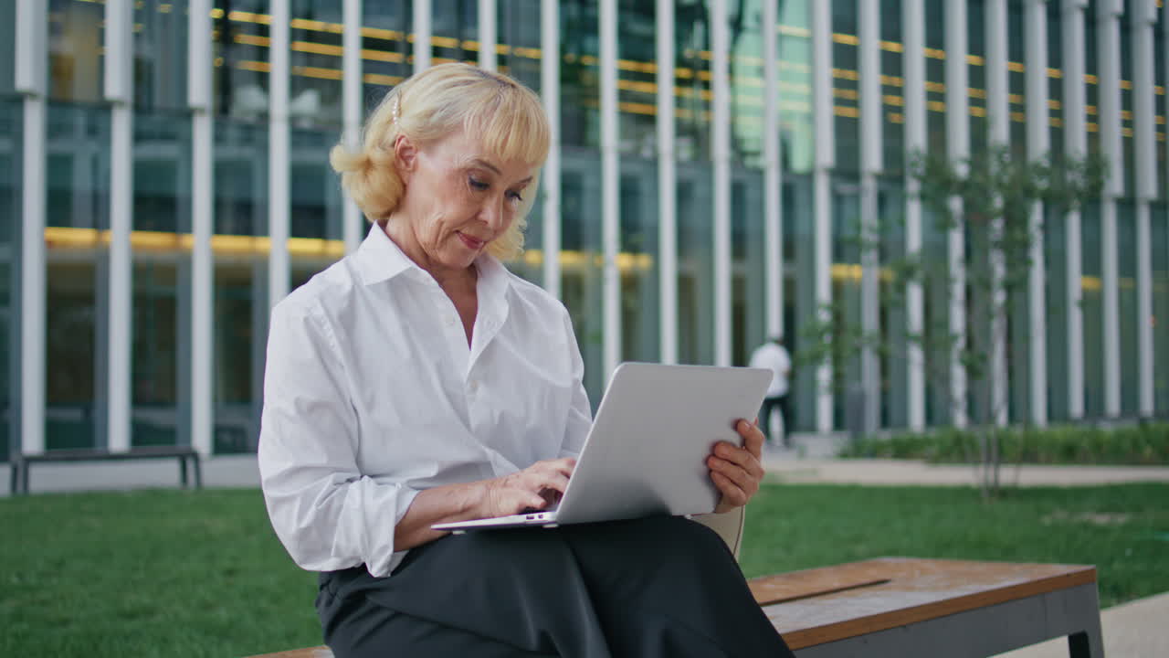 пожилая бизнесменка набирает текст на ноутбуке, сидя на уличной скамейке.